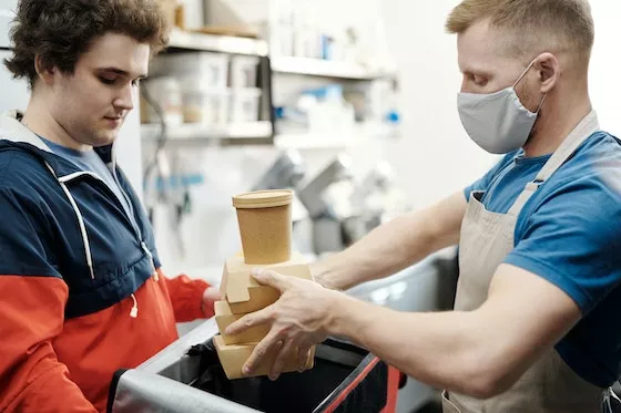 A dark-haired young man holds a cooler while another young man places food in to-go containers inside it